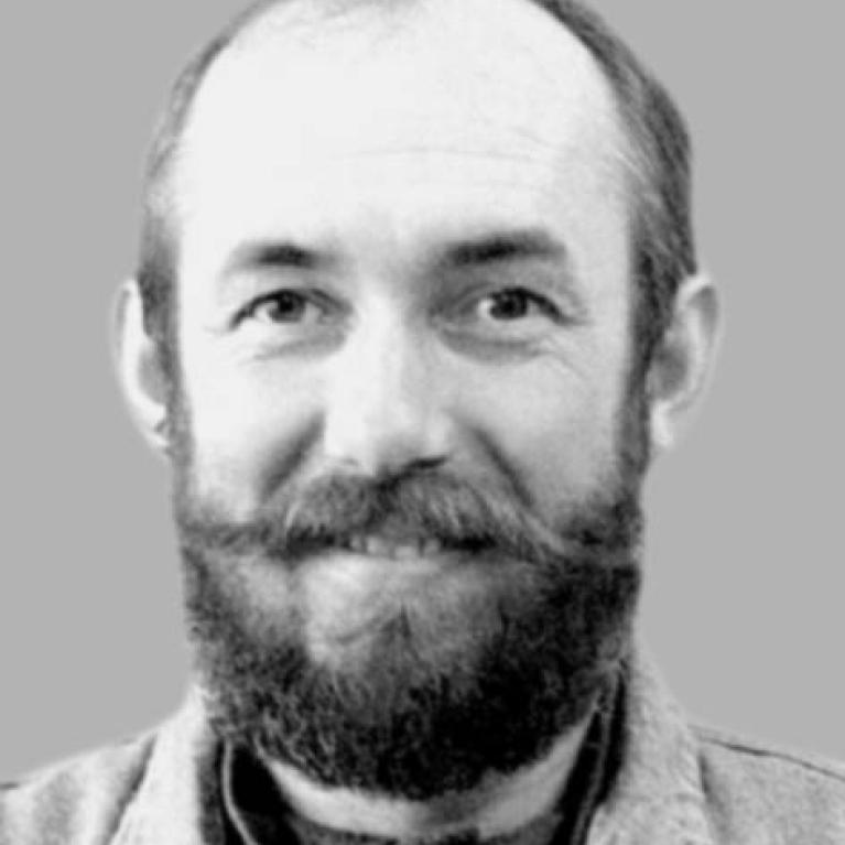 Black and white photograph of a person against a plain gray background. Visible features include a beard, short hair, and a light-colored jacket over a dark shirt.
