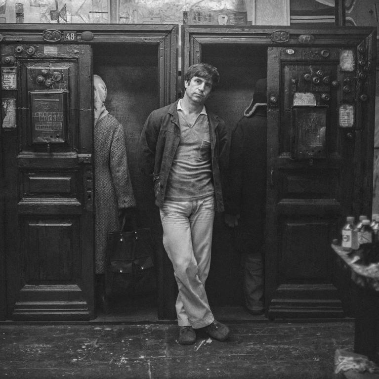 Black and white photograph showinga man leaning casually against the large-scale installation consisting of wooden doors and two human-size figures. 