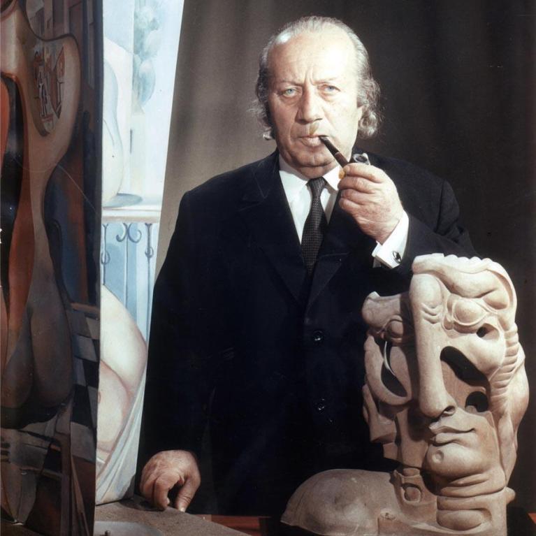 Photograph of a man in a black suit standing behind a table with a surrealist sculpture featuring multiple distorted faces and a painting with abstract shapes.
