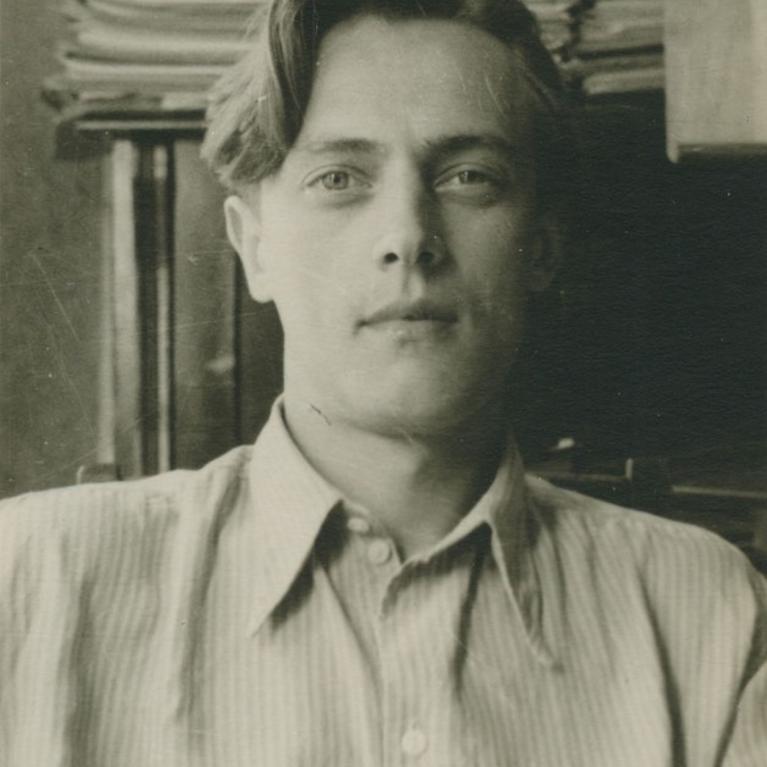 Black and white photograph of a person wearing a striped button-up shirt seated indoors with stacks of papers and a wooden piece of furniture in the background. The setting suggests an office or study environment, highlighting a mid-20th century style based on clothing and decor.
