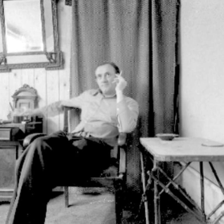 Black and white photograph showing a person sitting on a chair in a modest room with a small table and a cabinet. The setting includes a curtain, a framed mirror on the wall, and scattered items on the floor, suggesting a casual or contemplative moment.