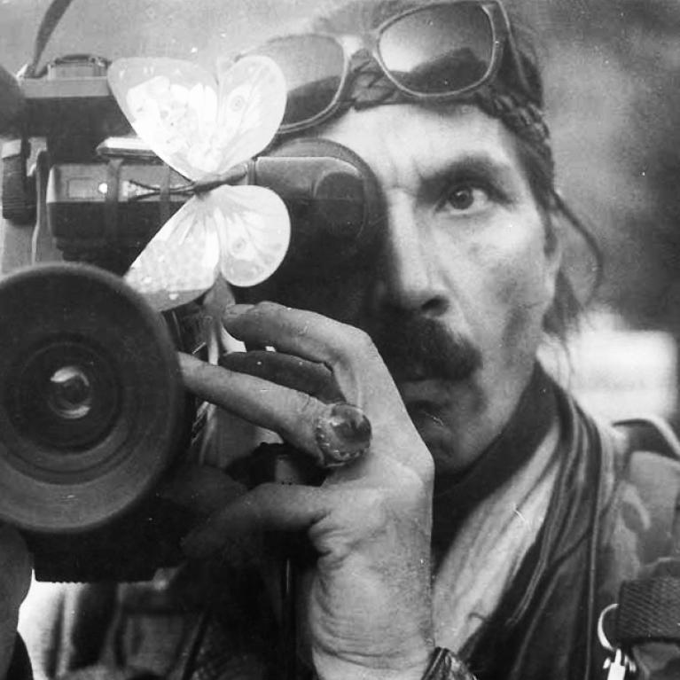 Black and white photograph of a person holding a video camera with a butterfly on it, capturing footage. The person wears sunglasses on his head and a jacket, suggesting a professional or documentary filming context.