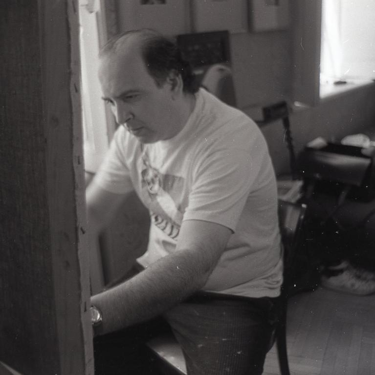 Black and white photograph showing a person seated and working on an easel painting. The setting appears to be an indoor room with a window, chairs, and various objects in the background, creating a casual and intimate atmosphere.