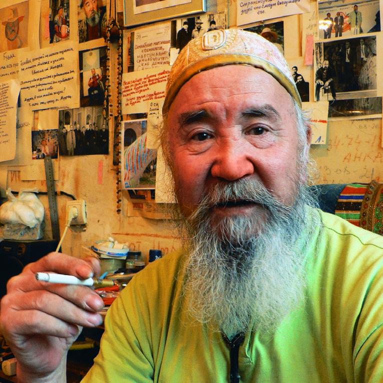 Photograph of a person with a long beard wearing a green shirt and holding a cigarette in a cluttered room filled with various photographs, handwritten notes, and decorations on the walls. The setting suggests a personal or creative space with a warm, lived-in atmosphere highlighted by colorful and detailed wall displays.