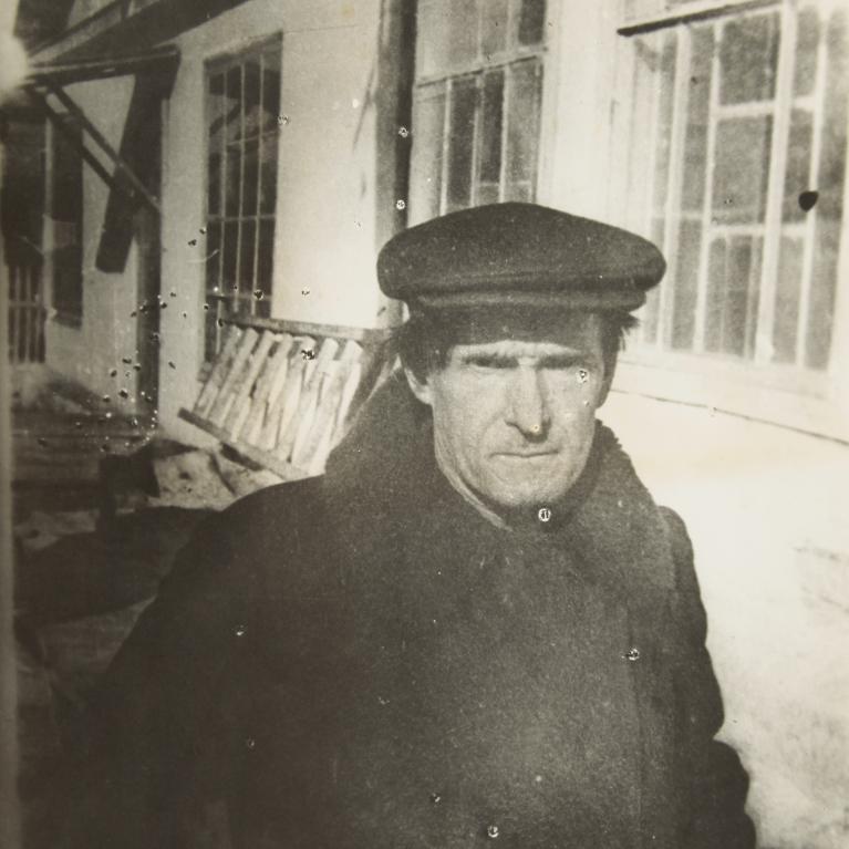 Black and white photograph showing a person wearing a dark coat and flat cap standing outside a building with large windows. The setting appears historical or vintage, with visible architectural details like window frames and a bench along the wall.