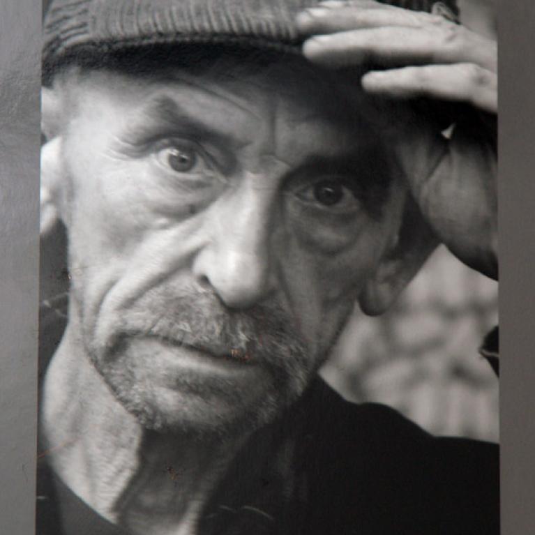 Black and white photograph showing a close-up of a person wearing a knit cap, with one hand raised to their forehead. 