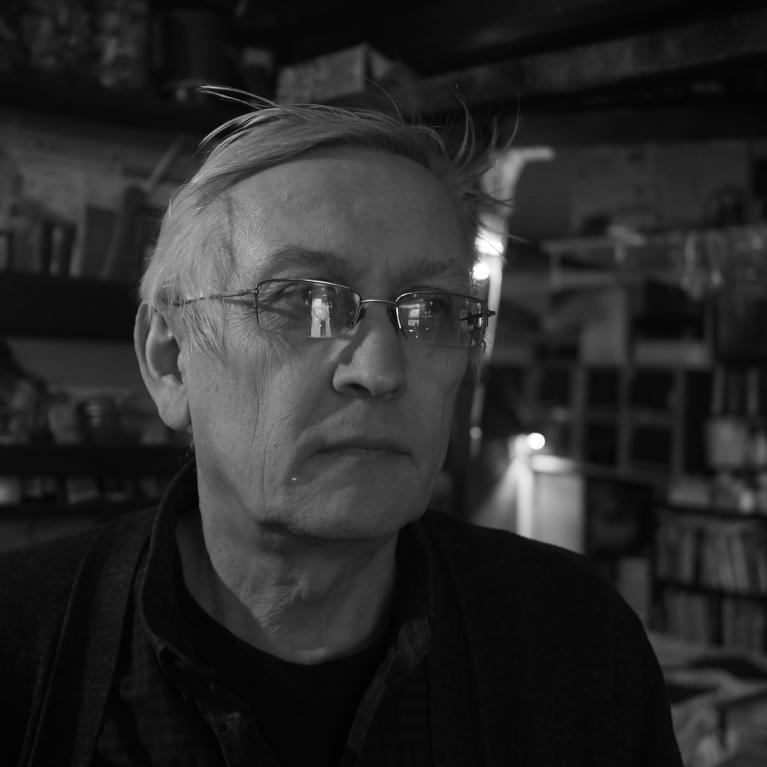 Black and white photograph of a person in an indoor setting filled with shelves and various objects. The scene highlights a cozy, cluttered environment suggesting a personal or work space.