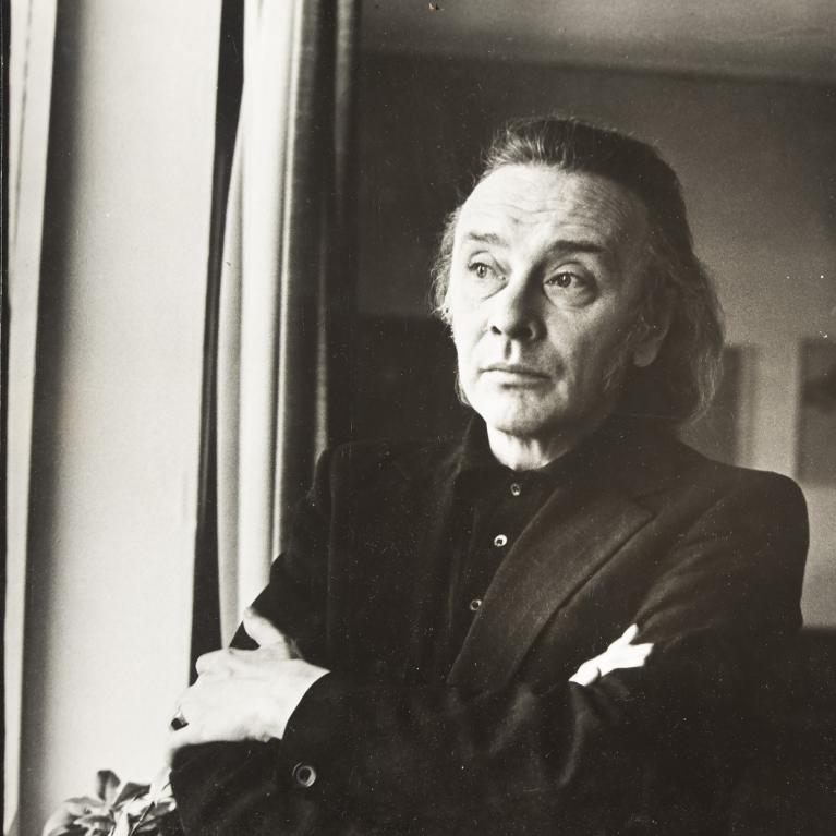 Black and white photograph of a person standing indoors near a window, wearing a dark suit jacket and a buttoned shirt. The person has arms crossed, with soft lighting creating contrast between the subject and background.