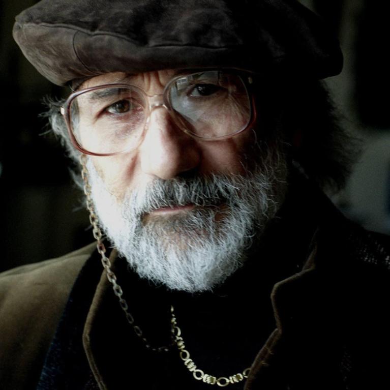 Photograph of a person wearing a brown flat cap, glasses, a dark jacket, and a gold chain necklace. The background is dimly lit, emphasizing the subject's attire and accessories.
