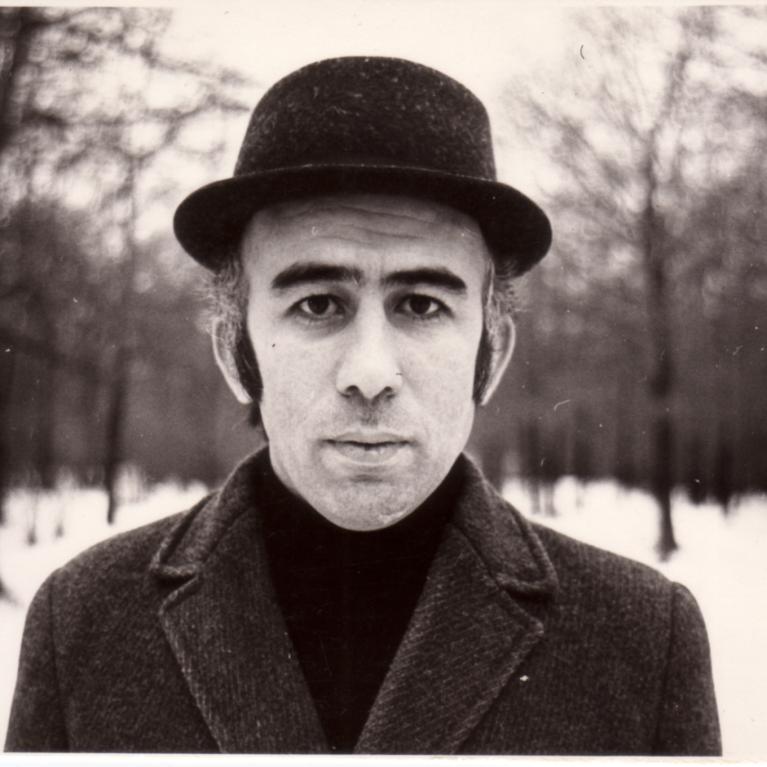 Black and white photograph of a person wearing a dark coat and bowler hat standing in a snowy forest with leafless trees.