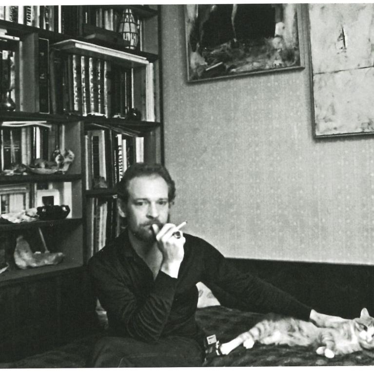 Black and white photograph showing a person sitting on a couch in a room filled with bookshelves and artwork on walls. The person is holding a cigarette and petting a cat lying on the couch, highlighting a relaxed, contemplative atmosphere.