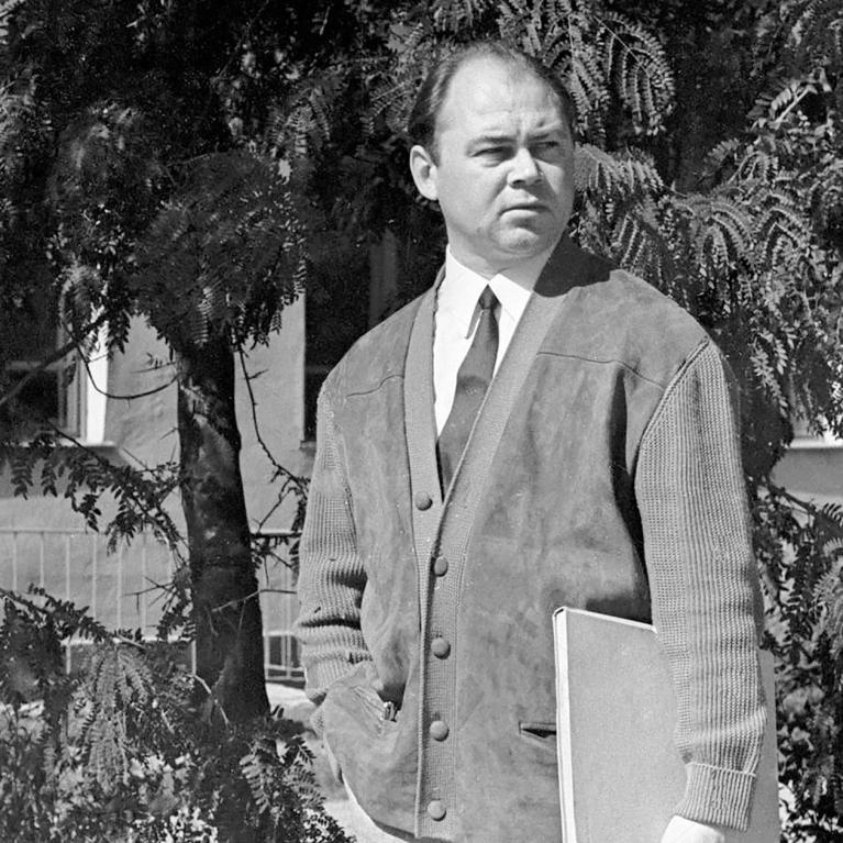 Black and white photograph of a person standing outdoors in front of trees and a building, wearing a buttoned cardigan over a shirt and tie, holding a folder or notebook in one hand. The setting suggests a casual or academic context with natural light casting shadows on the subject and surroundings.