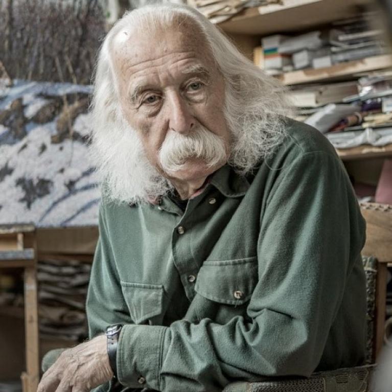 Photograph of an elderly person with long white hair and beard sitting in a cluttered room filled with stacks of papers and books. The person wears a green button-up shirt and a wristwatch, suggesting a setting of study or work in a personal or academic space.