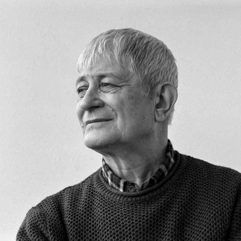 Photograph of an older man with short gray hair wearing a textured dark sweater over a plaid shirt, facing sideways against a plain light background. 