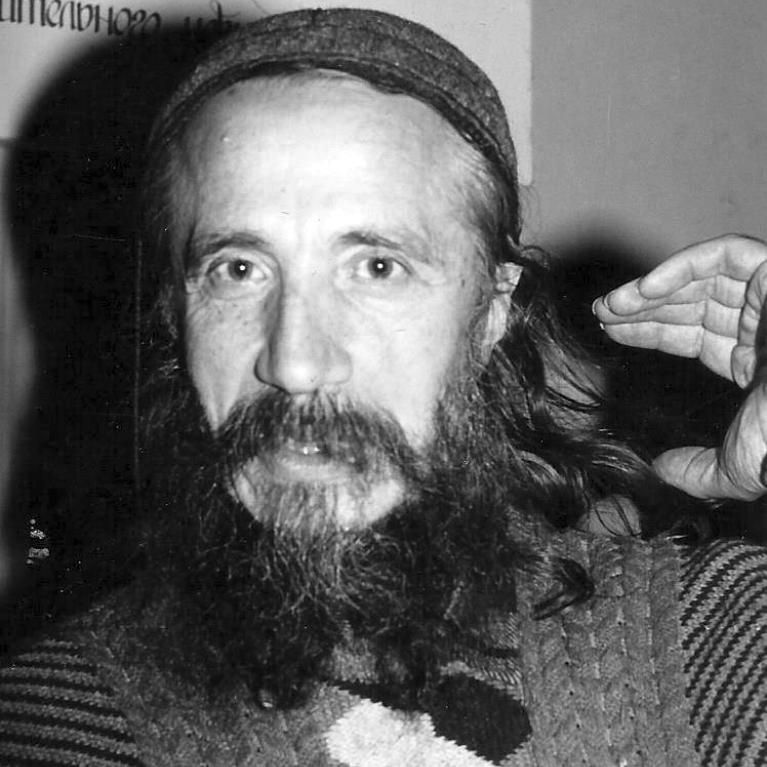 Black and white photograph of a person with long hair and a beard, wearing a patterned sweater and a hat, raising their right hand near their head. Background contains partially visible text in a decorative script.