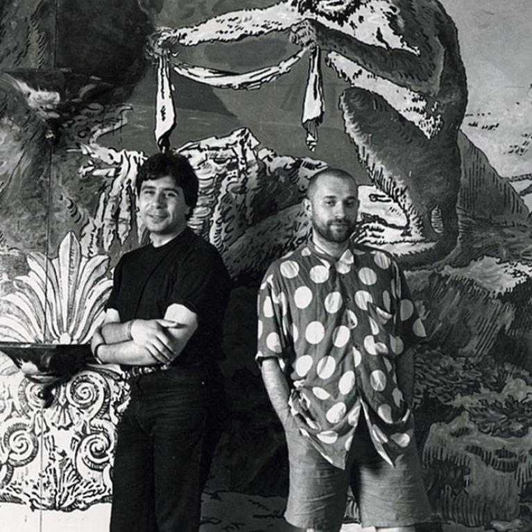 Black and white photograph of two men standing in front of a large mural depicting a mythical creature holding a cloth. One man wears a solid dark shirt and pants with arms crossed, while the other wears a polka dot shirt and shorts with hands in pockets.