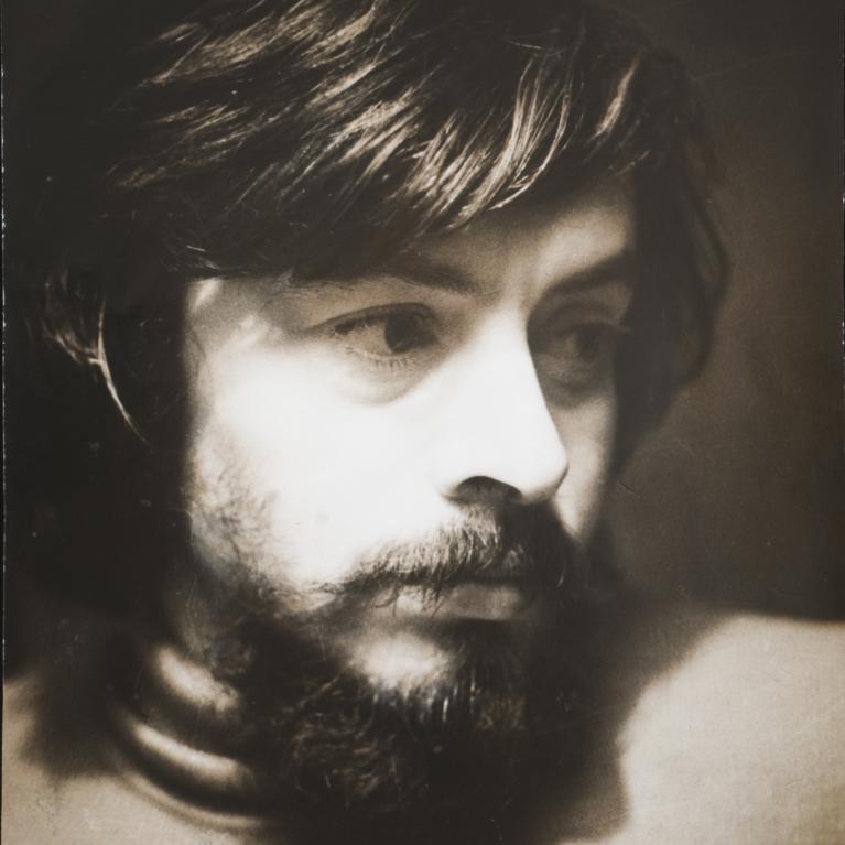 Photograph of a young man with medium-length dark hair and a full beard, looking contemplatively to the side. The sepia-toned portrait emphasizes soft lighting and shadow contrasts, highlighting facial features and texture of hair and beard.