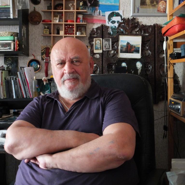 Photograph of an older man with a bald head and white beard sitting with arms crossed in a cluttered room filled with shelves, books, and various decorative items. Background includes framed pictures, a carved wooden panel, and electronic equipment, suggesting a personal or home office space.