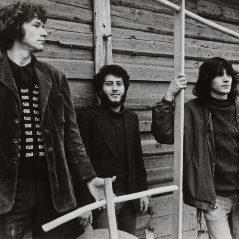 Black and white photograph of three young men standing in front of a wooden wall, each holding a wooden stick or pole. The image captures a casual, candid moment with varied clothing styles including a patterned sweater, jacket, and turtleneck.