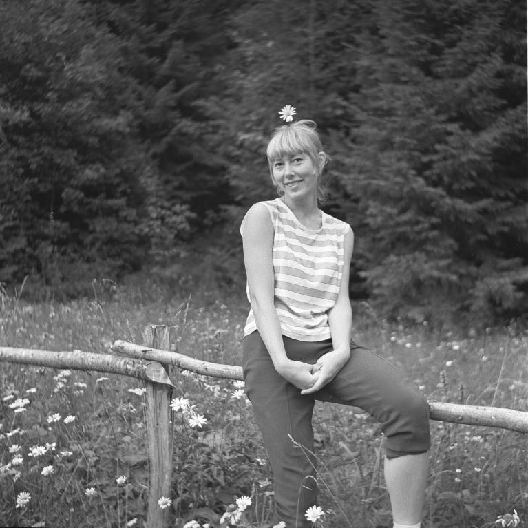 Black and white photograph of a woman sitting on a wooden fence in a field of wildflowers with a forest background. She wears a striped sleeveless top and cropped pants, smiling with a daisy tucked in her hair, conveying a relaxed, natural atmosphere.