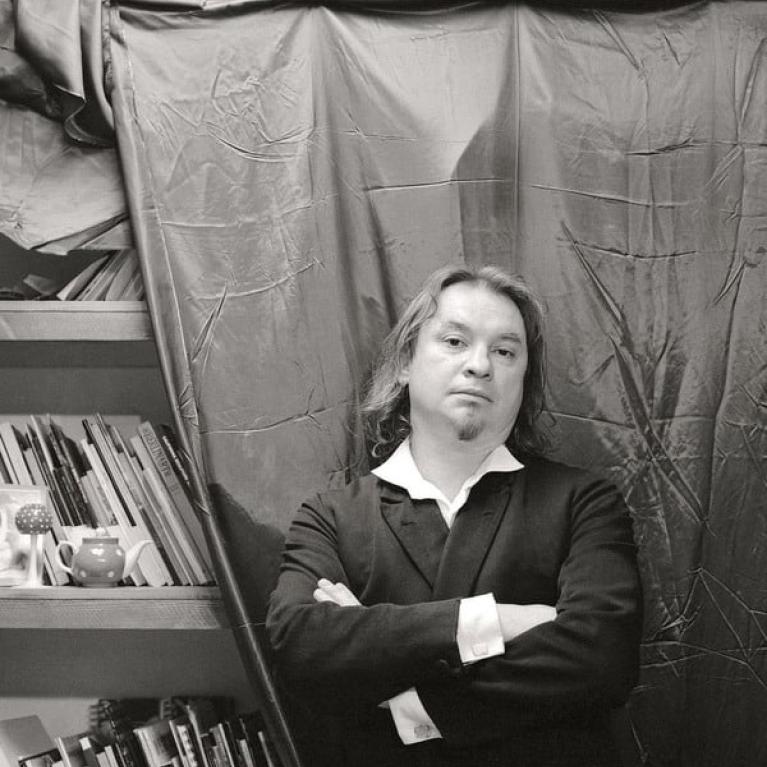 Photograph of a person standing with arms crossed in front of a draped fabric backdrop, wearing a dark suit and white shirt. Bookshelves filled with books and decorative items, including nested dolls, are visible on the left side, suggesting an indoor setting with a mix of formal and casual elements.