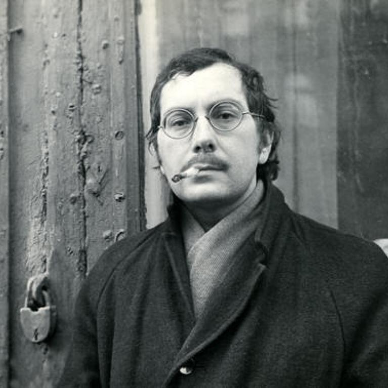 Black and white photograph of a man wearing round glasses, a dark coat, and a scarf, standing against a weathered wooden door. The man has a cigarette in his mouth and a neutral expression, with a vintage, mid-20th-century atmosphere.