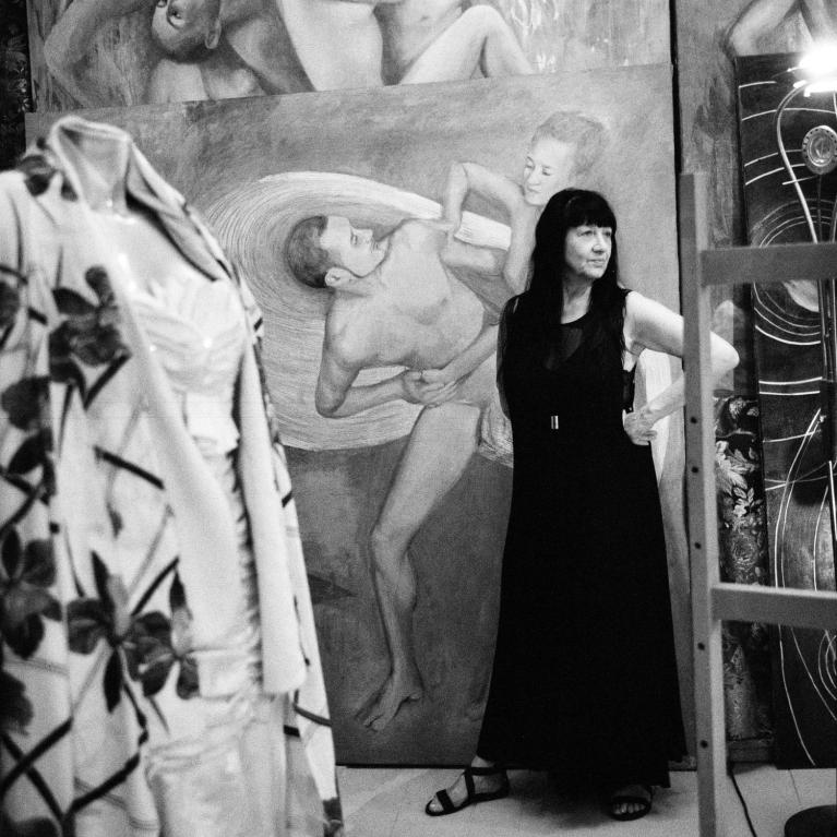 Black and white photograph of a woman standing in an art studio surrounded by large figurative paintings depicting intertwined human forms.