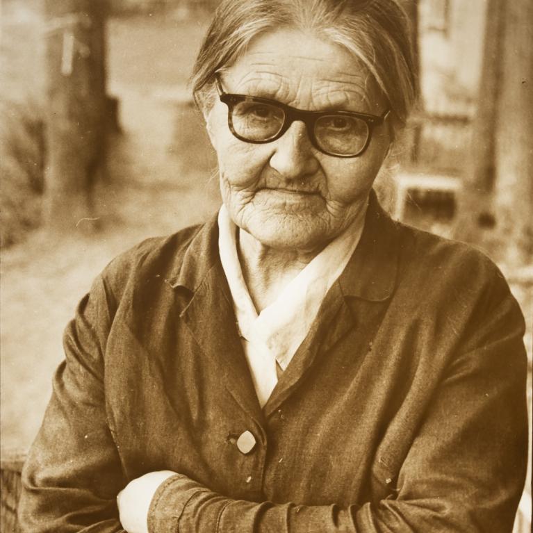 sepia-toned photograph of an older woman wearing glasses standing outside with her arms crossed