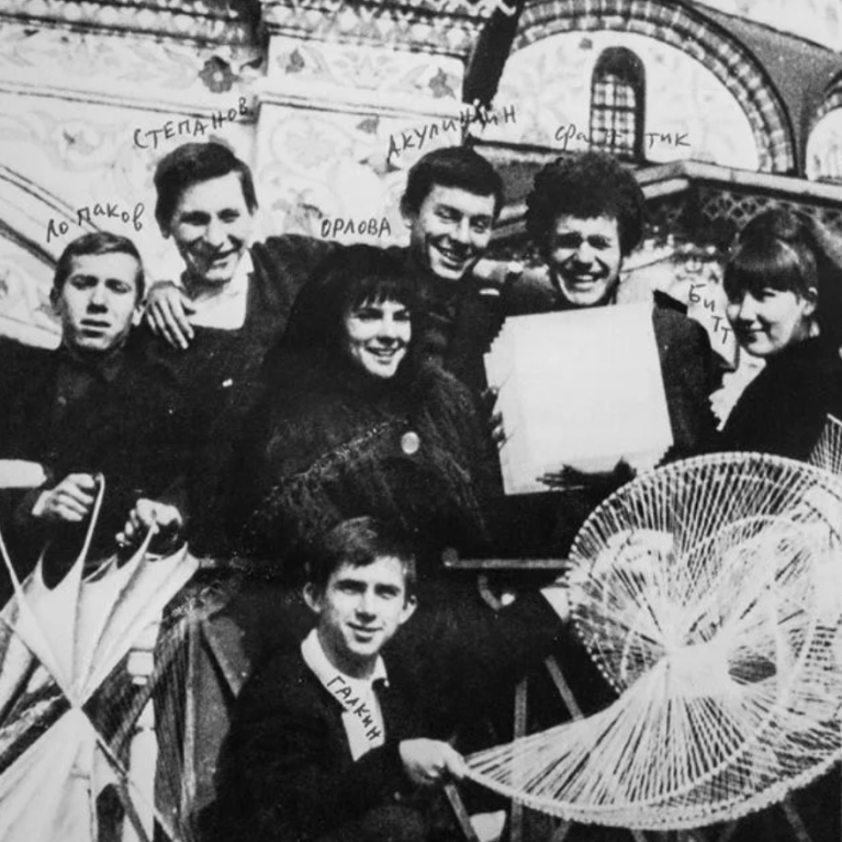 Black and white photograph of a group of people posing with various intricate geometric string art pieces in front of a decorated building facade. Handwritten names above each person identify individuals, highlighting a historical or cultural context.