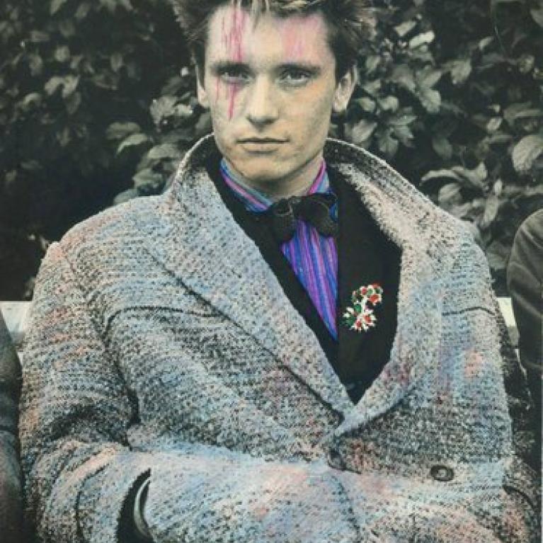 A photograph of a person sitting outdoors wearing a textured gray coat over a dark suit with a purple striped shirt and a black bow tie. The person has a floral brooch pinned to the suit and is positioned against a leafy background.