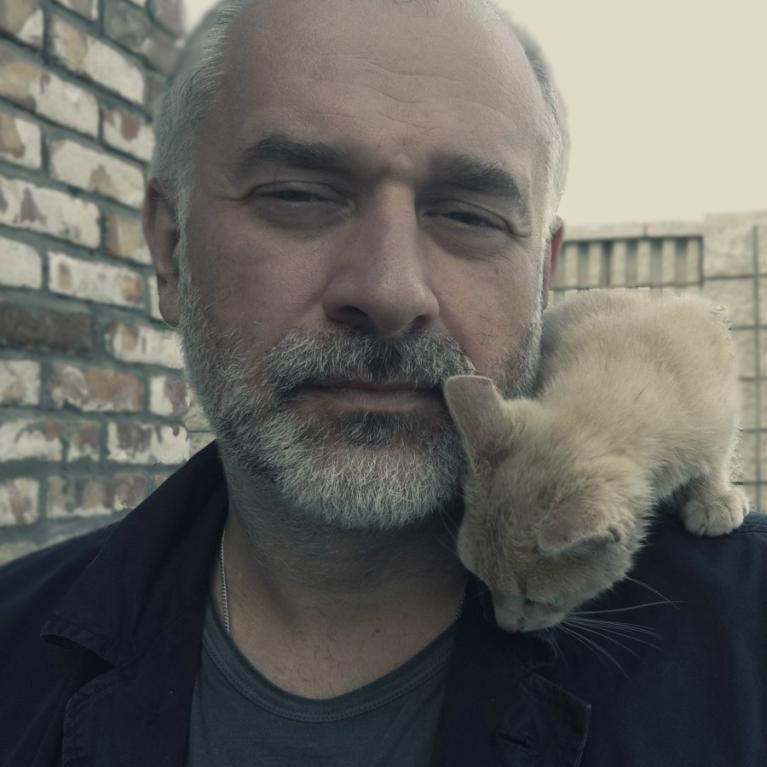 Photograph of a person wearing a dark jacket and gray shirt with a light-colored cat perched on their shoulder, nuzzling close to their neck. Background includes a brick wall on the left and a fence on the right, suggesting an outdoor setting.