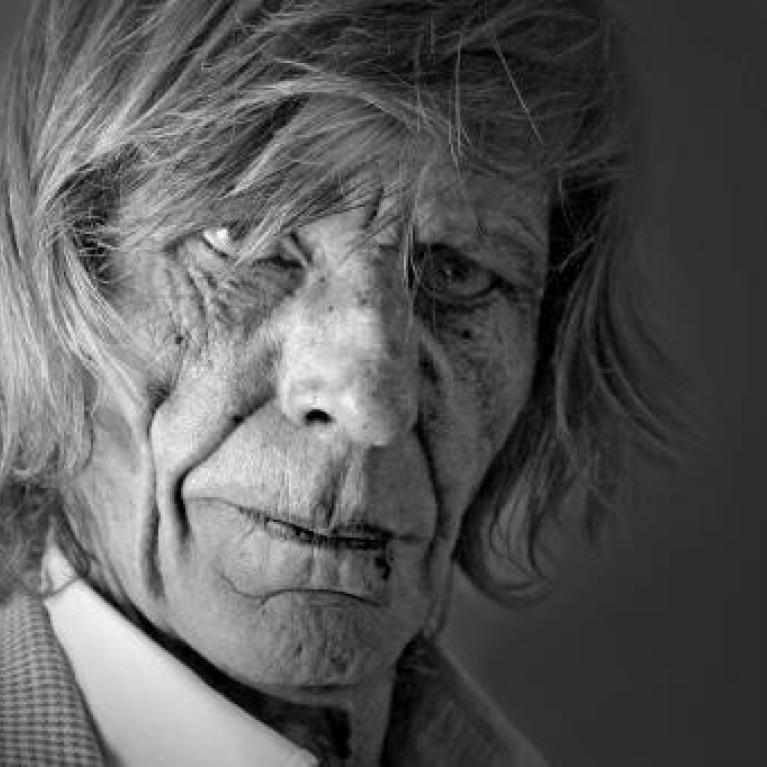 Black and white photograph of an elderly person with deep wrinkles, long hair, and a serious expression, wearing a checkered jacket and white shirt. The close-up portrait highlights texture and emotion, emphasizing age and character through lighting and contrast.