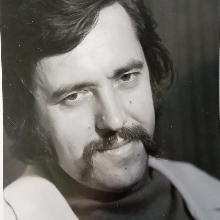 Black and white photograph of a man with dark hair, a thick mustache, and a slight smile, wearing a light-colored jacket over a dark shirt. The portrait captures a close-up view with a plain, dark background, emphasizing facial features and expression.