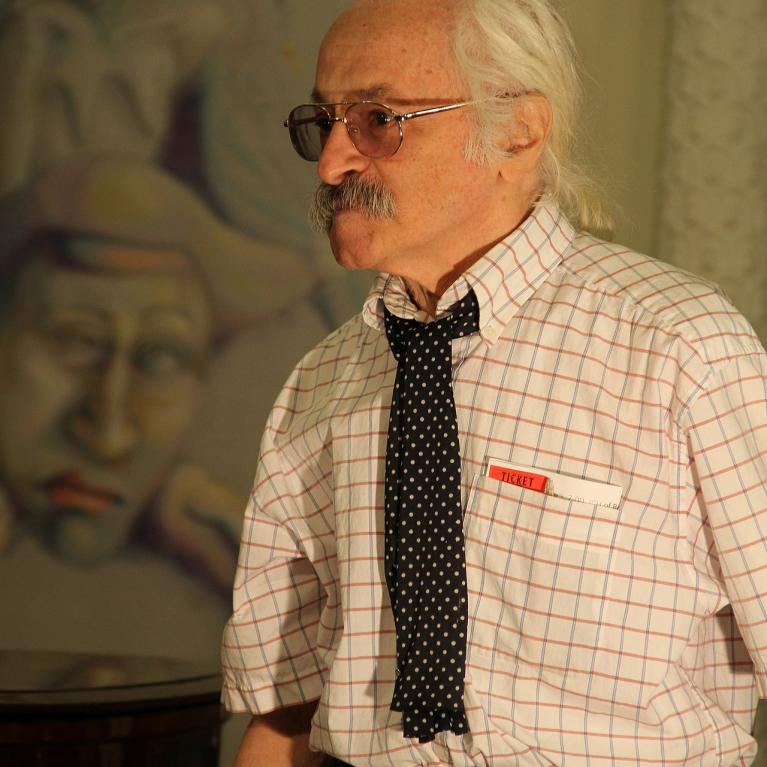 Photograph of an elderly man with white hair and mustache wearing glasses, a checkered shirt, and a black polka dot tie. Background features a blurred painting with abstract human faces and figures in muted colors.