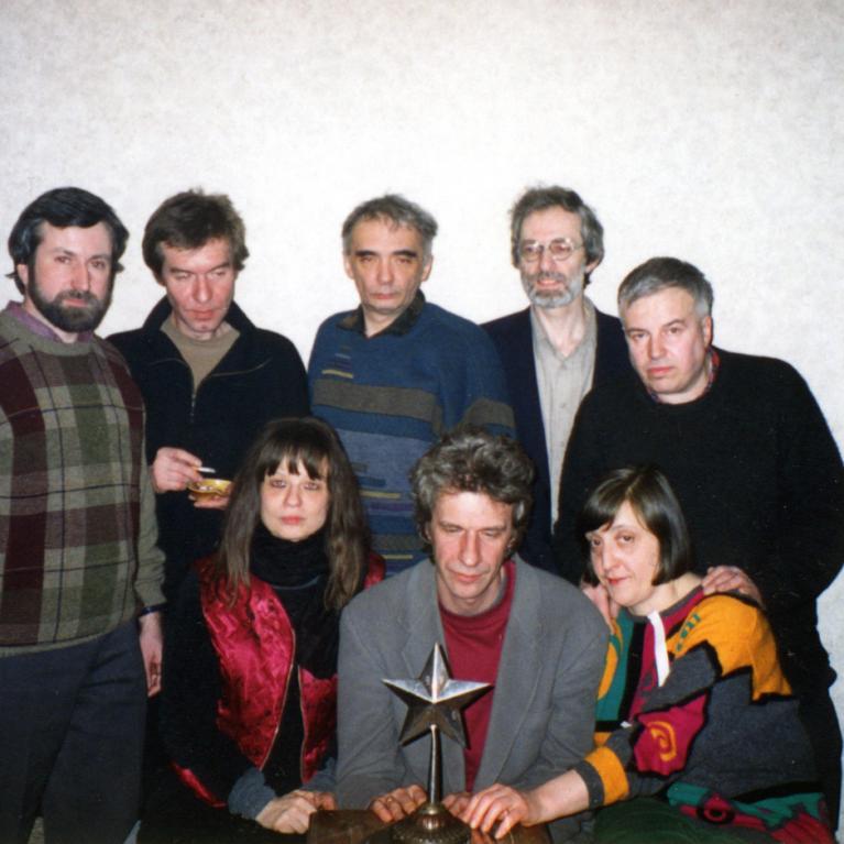 Photograph of a group of eight people, six men and two women, posing indoors against a plain wall with wooden furniture on either side. One man in front center holds a star-shaped trophy, while others stand or sit closely around him, dressed in casual sweaters and jackets.