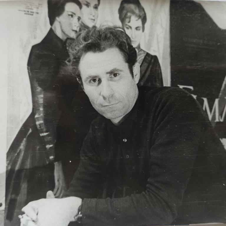 Black and white photograph of a man with curly hair wearing a dark sweater, leaning forward with hands clasped. Behind him are posters featuring three women and partially visible text, suggesting a setting related to theater.
