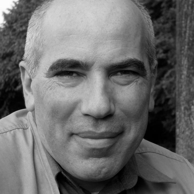 Photograph of a middle-aged man with short gray hair and a slight smile, wearing a collared shirt and jacket. Background features blurred trees, suggesting an outdoor setting.