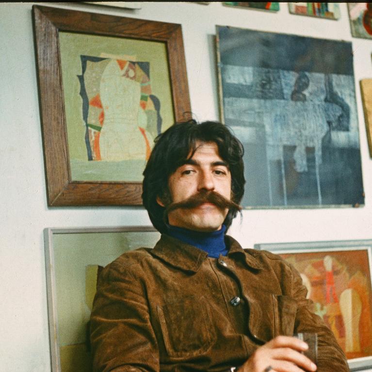Photograph of a man with a thick mustache and brown jacket sitting indoors against a wall adorned with framed abstract artwork. The setting suggests a casual, artistic environment with warm lighting and a relaxed atmosphere.