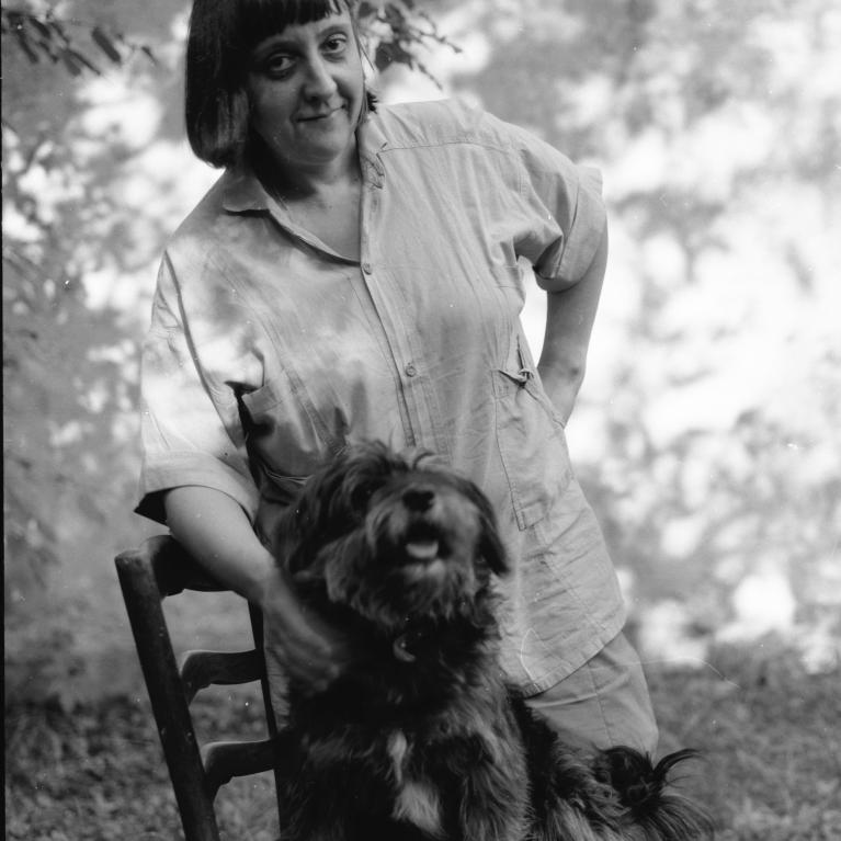 Black and white photograph of a woman standing behind a wooden chair with a small, fluffy dog sitting on it. The woman is smiling slightly, wearing a loose shirt, with leafy branches and a blurred outdoor background.