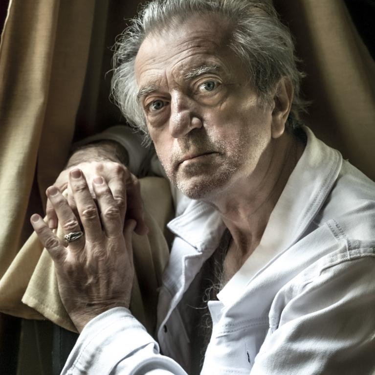 Photograph of an elderly man with gray hair and a contemplative expression, wearing a white shirt and resting his hands on his knee near beige curtains. The lighting highlights facial wrinkles and texture, emphasizing a reflective or somber mood.