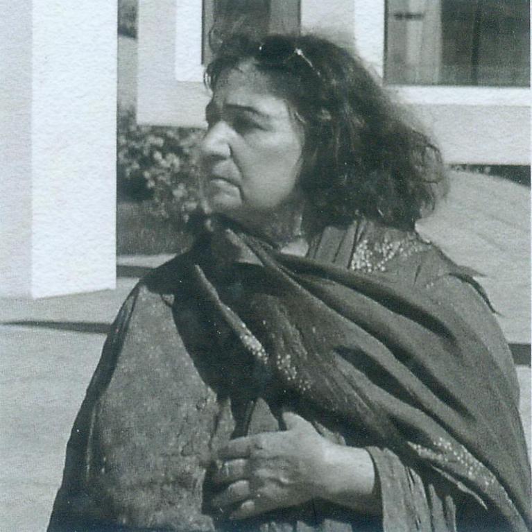 Black and white photograph of a woman with curly hair wearing a patterned shawl, looking to her left. Background includes a building with large windows and some foliage.