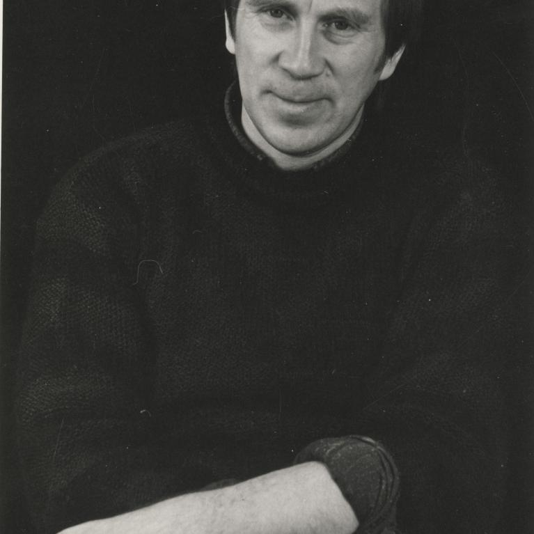 Photograph of a man sitting with arms crossed, wearing a dark sweater and looking slightly to the side with a subtle smile. The black-and-white portrait features a plain dark background, emphasizing the subject's facial expression and relaxed posture.