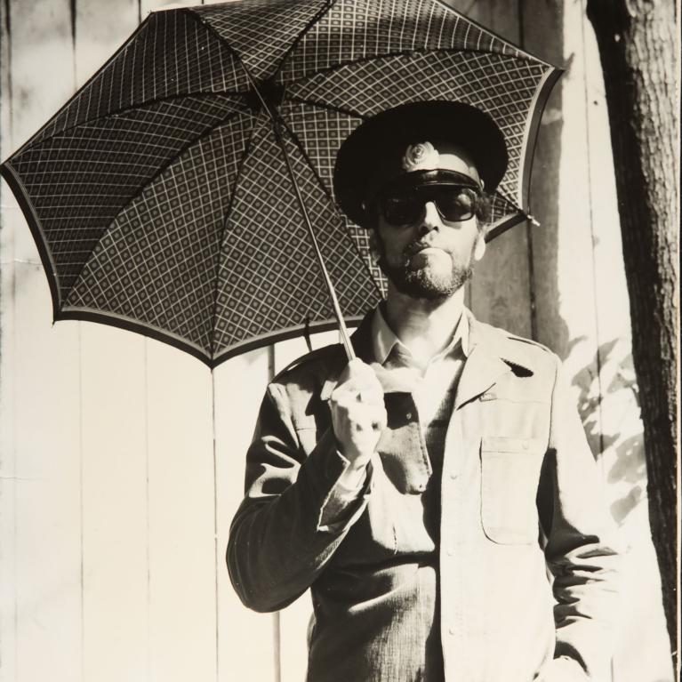 Black and white photograph of a man wearing a military-style jacket, cap, and sunglasses holding a patterned umbrella. The man stands against a wooden fence with sunlight casting shadows, creating a contrast between light and dark areas.