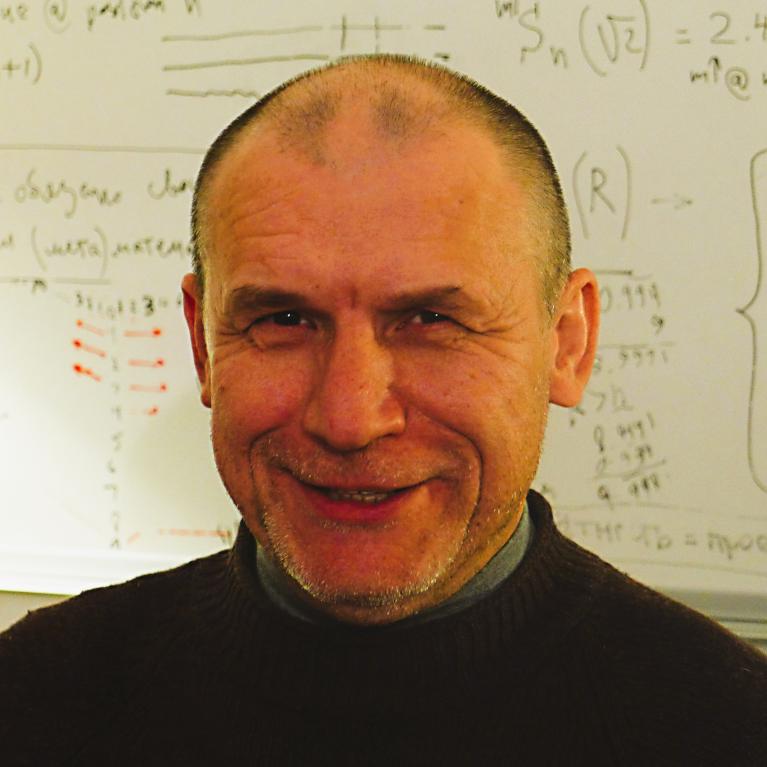 Photograph of a middle-aged man smiling in front of a whiteboard covered with mathematical equations and diagrams. 