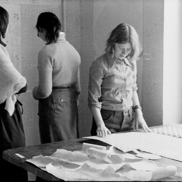 Black and white photograph showing three women in a room with a large table covered in papers and fabric samples. One woman is closely examining a large sheet on the table