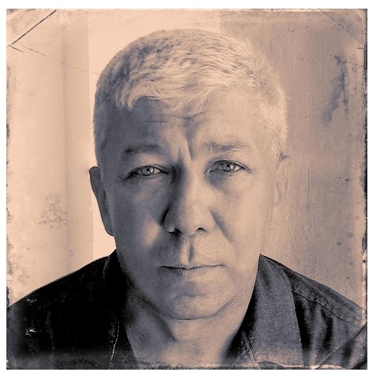 Photograph of a middle-aged man with short, light-colored hair and a serious expression, facing forward against a plain, textured background. The photo has a vintage, sepia-toned filter with visible wear marks and scratches around edges, giving an aged appearance.