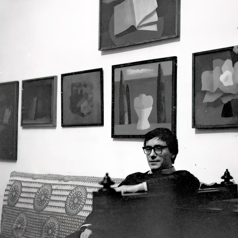 Black and white photograph of a man wearing glasses sitting on a patterned couch in front of a wall displaying six abstract paintings. Paintings feature geometric and floral shapes with dark backgrounds, arranged in two rows, highlighting a modern art gallery or studio setting.