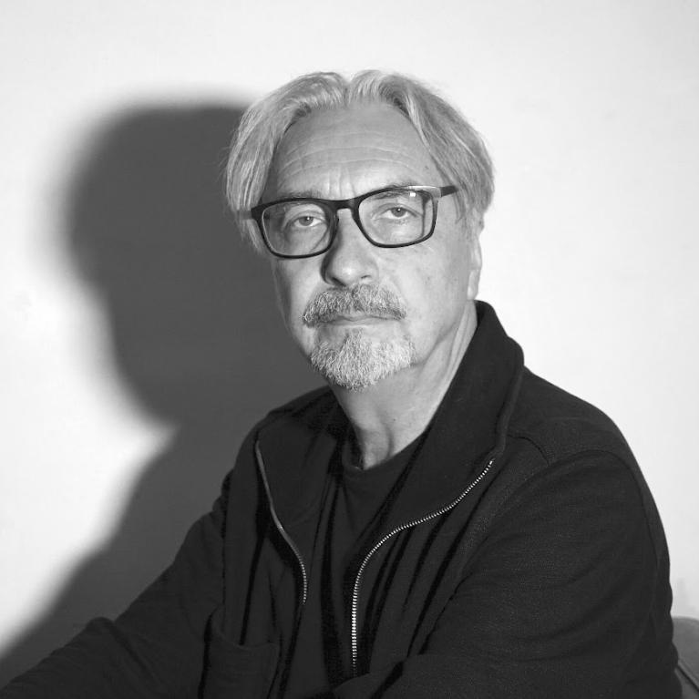 Black and white photograph of an older man with gray hair, glasses, and a mustache wearing a dark zip-up jacket and shirt. The man is seated against a plain background with a distinct shadow cast on the wall behind him.