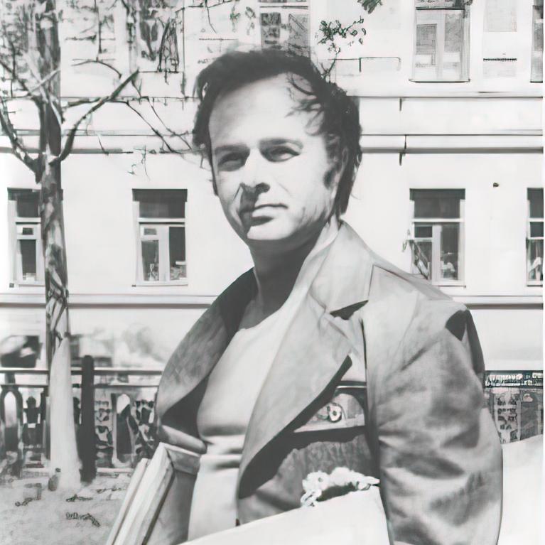 Black and white photograph of a man standing outdoors in front of a building, holding books and papers. The man wears a jacket over a shirt, with a tree and windows visible in the background, suggesting an urban or academic setting.