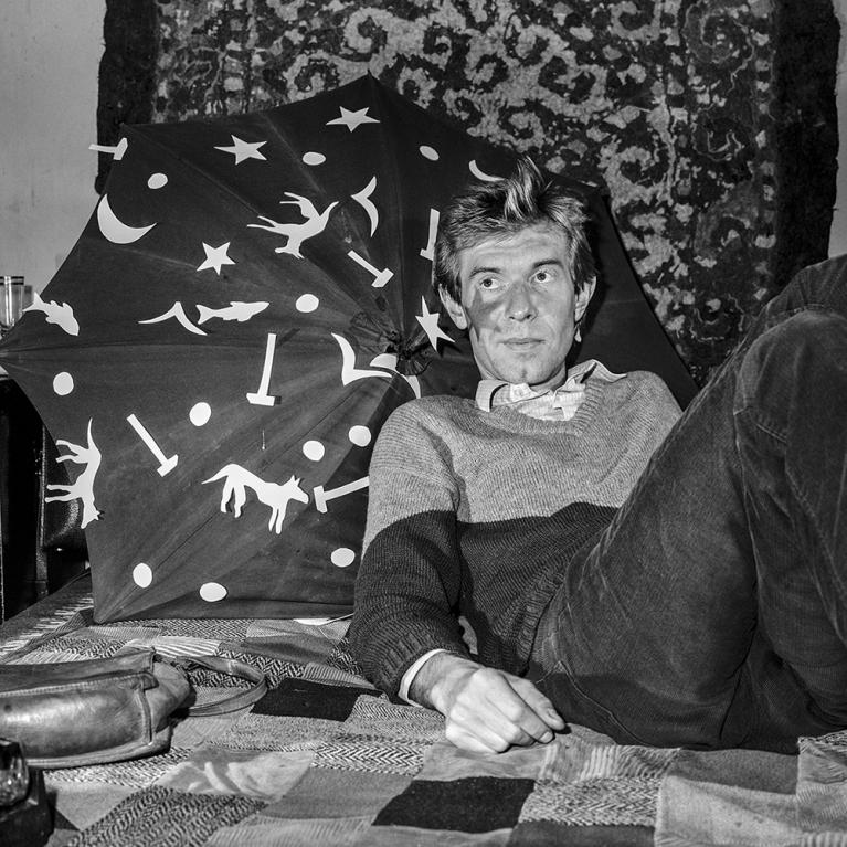 Black and white photograph of a man reclining on a patterned quilt with a decorative umbrella featuring stars, moons, and animal silhouettes behind him. The setting appears casual and indoors, with textured wall hanging and personal items like a bag and bottle visible, suggesting a relaxed, intimate environment.
